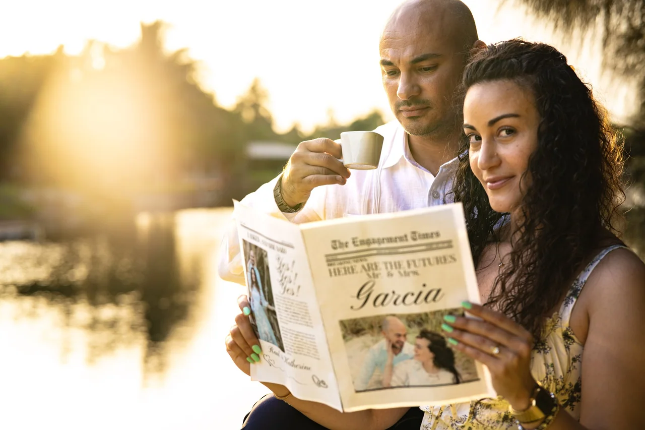 An engaged couple reading a personalized newspaper beside a lake in warm evening light.