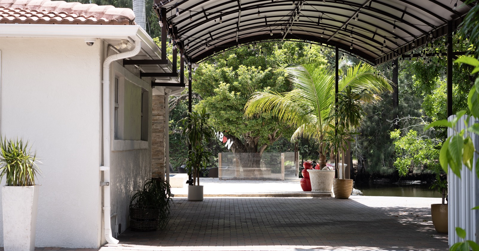 A long arched canopy shelters an open passage between a building and a wooded waterside patio.