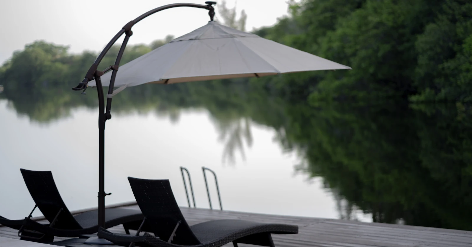 A cantilevered patio umbrella supported by one curved mast shades two empty chairs beside calm water.